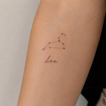 Minimalistické tetování jména „Leo“ v tenkém rukopisném písmu na vnitřní straně předloktí, nad ním jemně tečkami a linkami vyznačené souhvězdí Lva – ukázka spojení jména se symbolem zvěrokruhu.