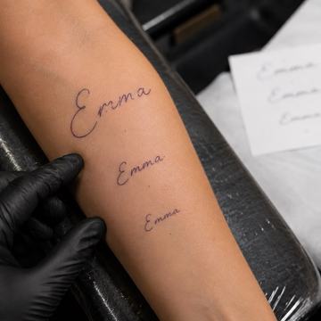 Předloktí s třemi variantami nápisu „Emma“ v tenkém rukopisném písmu nad sebou; tatér v černé rukavici ukazuje na největší návrh, vedle leží papírová předloha – ukázka volby velikosti a umístění jmenného tetování na ruku.