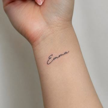 Jemné tetování jména „Emma“ na vnitřní straně zápěstí, tenké rukopisné písmo – minimalistická a dobře čitelná ukázka jmenného motivu na zápěstí.
