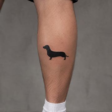 Malé pánské tetování na lýtku: minimalistická blackwork silueta jezevčíka umístěná nad kotníkem, dobře viditelná při nošení kraťasů – ukázka nenápadného motivu na nohu.