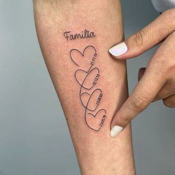 Jemné rodinné tetování na předloktí: nápis „Familia“ a čtyři propojená srdce s daty narození, minimalistická linka; prst ukazuje na motiv.