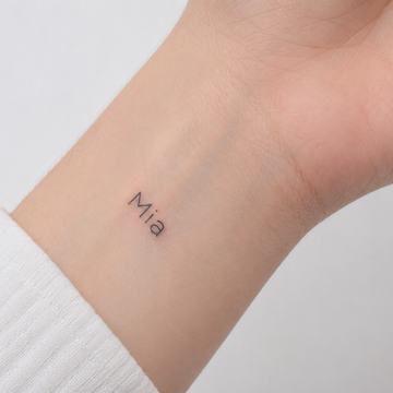 Minimalistické tetování jména „Mia“ malým čistým písmem na vnitřní straně zápěstí.