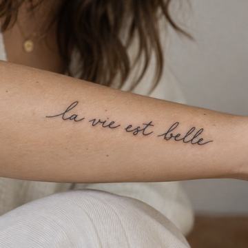 Minimalistický tetovací nápis „la vie est belle“ (francouzsky „Život je krásný“) jemným rukopisným písmem na vnitřní straně předloktí – příklad krátkého citátu s důrazem na čitelnost a vhodné umístění.
