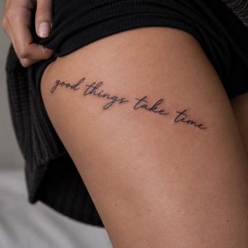 Jemný nápis „good things take time“ (Dobré věci potřebují čas) rukopisným písmem vytetovaný na horní části stehna; příklad krátkého anglického motta s důrazem na čitelnost a vhodné umístění.