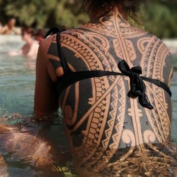 Rozsáhlé černé tribal tetování přes záda a ramena: silné linky, souvislé plochy a negativní prostor; ornamentální, částečně symetrická kompozice plynule kopíruje anatomii. Foceno zezadu ve vodě.