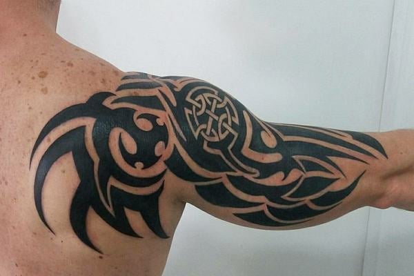 Velké černé tribal tetování přes rameno a horní paži: silné linky, souvislé plochy a negativní prostor, ornamentální uzel na rameni; kompozice kopíruje anatomii a působí jako polorukáv.