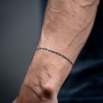 Minimalistické pánské tetování na zápěstí: jemná černá řetízková linka obepínající ruku, čisté provedení jemnými linkami, decentní motiv.