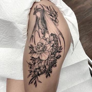Blackwork tetování evropského draka s roztaženými křídly propleteného s pivoňkami a drobnými květy na vnější straně stehna; čisté linie, jemné stínování a kompozice kopírující anatomii.