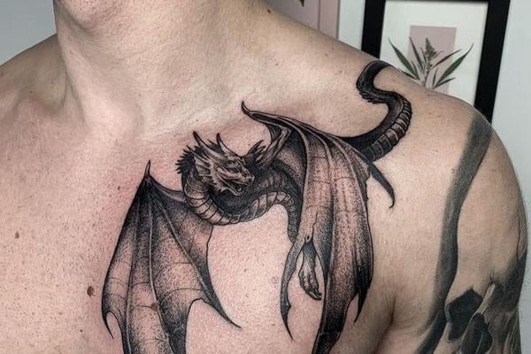 Blackwork tetování evropského draka s roztaženými křídly a dlouhým ocasem přes klíční kost, horní část hrudi a rameno; 