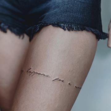 Jemné tetování nápisu tenkou černou linkou na horním stehně – minimalistická, horizontální kompozice jako inspirace pro tetování na nohu, snadno skrytelná oblečením.