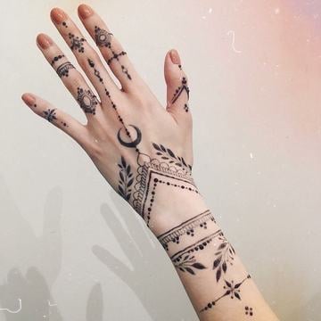 Jemné dámské tetování na prstech a hřbetu ruky: fine line a dotwork s prstýnkovými pásky, půlměsícem, listy a geometrickými ornamenty navazujícími až na zápěstí.