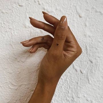 Jemné tetování na prstech a hřbetu ruky ve stylu fine line/dotwork: had, geometrická hvězdice, půlměsíc a drobné tečky s linkami mezi klouby; stříbrné prsteny podtrhují dojem tetování jako decentního šperku.