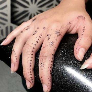 Jemné dámské tetování na prstech a hřbetu ruky: minimalistické fine line a dotwork s hvězdami, tečkami a romby, rozložené na více prstech do prstýnkových pásků a geometrických ornamentů.