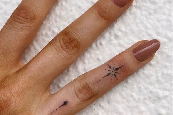 Ruka s jemným tetováním na prsteníčku: fine line/dotwork hvězda se jiskrami mezi klouby a drobné tečky s linkou po boku prstu jako decentní prstýnkový motiv; nude manikúra, jeden nehet třpytivý.
