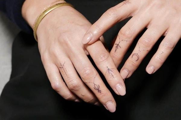 Dvě dámské ruce s jemným tetováním na prstech: minimalistické fine line symboly (měsíc, hvězdičky, tečky, linky a drobné znaky) rozmístěné mezi klouby a po stranách prstů jako prstýnkové pásky; na zápěstí jednoduchý zlatý náramek.