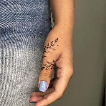 Jemné tetování na boční straně palce – minimalistická větvička s lístky ve fine line stylu, vedená od hřbetu ruky podél prstu jako decentní prstýnkový detail.