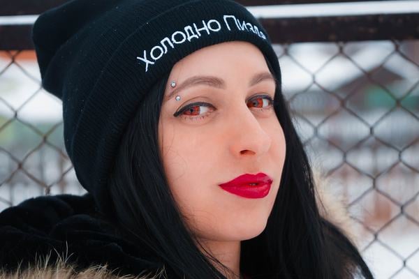 Osoba venku v čepici s vertikálním piercingem ve vnější části obočí (zahnutá činka); ukázka moderního vzhledu a správného umístění piercingu.