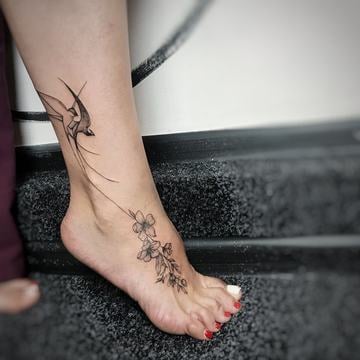 Jemné dámské tetování na kotníku: linework vlaštovka na vnější straně kotníku propojená tenkou linií s květinovou větvičkou, která pokračuje přes nárt k prstům – minimalistický motiv, který kopíruje tvar nohy.