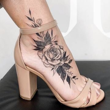 Dámské tetování na kotníku – černobílá růže s listy v linework s jemným stínováním, motiv sahá od nártu přes kotníkovou kost směrem k lýtku; 