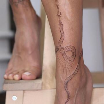 Jemné linework tetování hada na vnější straně ženského kotníku, protažené podél kotníkové kosti směrem k lýtku, s dekorativním dotworkem a motivem půlměsíce se šipkou nad hlavou – ukázka minimalistického motivu na kotník.
