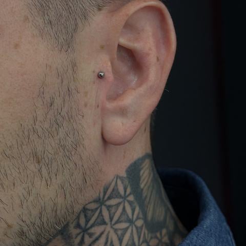 Minimalistický piercing v ušním lalůčku s drobným tmavým šperkem působí decentně, ale zároveň osobitě. Jednoduchý design vytváří zajímavý kontrast s výrazným geometrickým tetováním na krku.