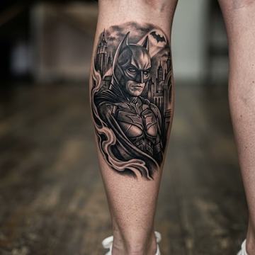 Pánské tetování na lýtku: vertikální černošedý realismus/blackwork s Batmanem, panoramatem města a bat‑signálem, přizpůsobené tvaru svalu.