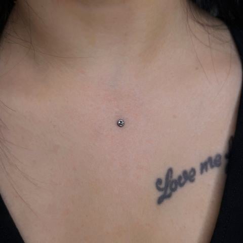 Minimalistický mikrodermální piercing v oblasti dekoltu s jediným třpytivým kamínkem. Drobné zdobení na hladké pokožce vytváří elegantní akcent, který vkusně doplňuje tetovaný nápis u klíční kosti.