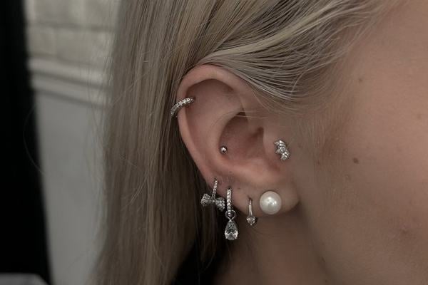 Detail ucha s tragus piercingem – malý, nenápadný labret v chrupavce před vstupem do zvukovodu; ucho má také helix kroužek a několik náušnic v lalůčku (perla a visací kamínky), což ukazuje tragus jako decentní doplněk vícenásobného piercingu.