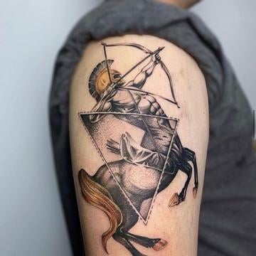 Černošedé realistické tetování na horní paži: mytologický kentaur-lučištník v helmě napínající luk, kompozice s geometrickým trojúhelníkem a dotwork texturou; čisté linky a precizní stínování jako výrazný motiv na ruku.