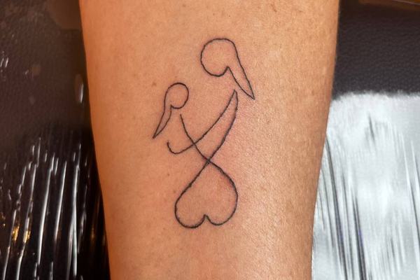Jemné rodinné tetování na předloktí: minimalistická linka dvou ptáků s propletenými krky končící srdcem – decentní symbol blízkosti a soudržnosti rodiny.