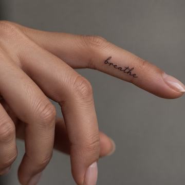 Minimalistický tetovací nápis „breathe“ (Dýchej) drobným rukopisným písmem podél boku prstu – příklad krátkého anglického motta s důrazem na čitelnost a vhodné umístění.