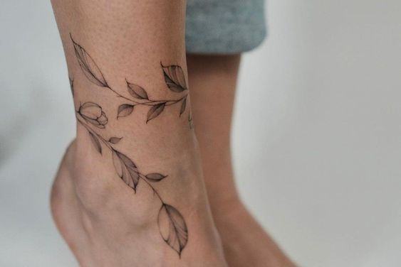 Jemné černé linework tetování větvičky s listy, které obepíná ženský kotník jako náramek a lehce pokračuje k lýtku – minimalistická ukázka tetování na kotník.