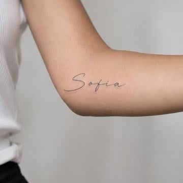 Jemné tetování jména „Sofia“ v tenkém rukopisném písmu, vodorovně na vnitřní straně paže nad loktem – ukázka minimalistického, čistě čitelného jmenného motivu.