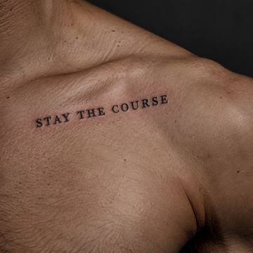 Minimalistický nápis „STAY THE COURSE“ tetovaný podél klíční kosti, verzálkové serifové písmo – příklad krátkého anglického motta s důrazem na čitelnost a vhodné umístění.