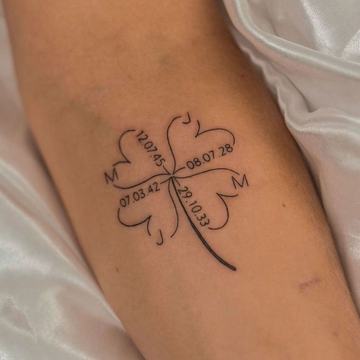 Jemné rodinné tetování tenkou linkou: čtyřlístek ze srdcí s iniciálami M a J a čtyřmi daty narození – minimalistický symbol rodiny, blízkosti a štěstí.