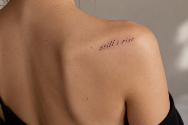 Minimalistický tetovací nápis „still i rise“ malými písmeny jemným drobným písmem nad lopatkou u ramene – příklad krátkého anglického motta s důrazem na čitelnost a vhodné umístění.