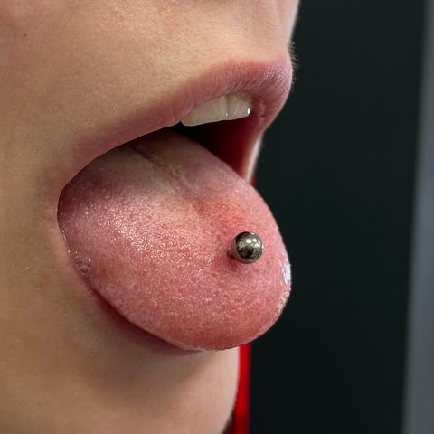 Piercing jazyka s tmavým šperkem vytváří odvážný a výrazný detail. Kontrastní kulička dodává výsledku hravý a nekonvenční charakter.