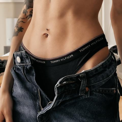 Piercing pupíku zde působí jako jemný šperk zvýrazňující pevnou linii břicha. Lesklý detail kontrastuje s tmavým outfitem a dodává celku moderní, smyslnou energii.